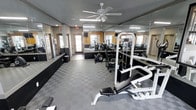 This single-story fitness center offers a functional and well-organized workout environment designed to accommodate a variety of exercise needs. The space features a checkered gray rubber flooring that provides both durability and traction, making it well-suited for high-traffic use. Large mirrors line multiple walls, creating a sense of openness while allowing users to monitor their form during workouts.

The room is outfitted with a selection of cardio equipment, including elliptical machines, as well as strength training equipment such as a weight bench and cable machine. A ceiling fan mounted at the center of the room aids in air circulation, complementing the fluorescent overhead lighting that ensures the space is brightly and evenly lit. Natural light also filters in through windows along one wall, adding to the overall brightness of the environment.

The walls feature a warm yellow-toned accent that contrasts with the gray tones of the floor and equipment, contributing to an energizing atmosphere. Emergency exit signage is clearly visible, and the layout allows for comfortable movement between stations. Overall, the space is practical, clean, and conducive to an effective workout experience.