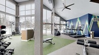 Located at 7301 Bristol Village Dr in Bloomington, Minnesota, this well-appointed fitness center offers a spacious, single-floor layout designed to accommodate a comprehensive range of workout activities. The space is defined by its dramatic vaulted ceiling with exposed wood beam accents and a large ceiling fan, creating an open and airy atmosphere. Floor-to-ceiling windows span a significant portion of the exterior walls, flooding the interior with natural light and offering views of the surrounding outdoor landscape.

The fitness center is divided into distinct functional zones. The cardio area features multiple treadmills and elliptical machines lined along a boldly accented blue accent wall with geometric artwork. A dedicated free weights section includes a full dumbbell rack, kettlebells, barbells, and adjustable benches positioned on green turf flooring, which contrasts with the neutral carpet in the cardio zone. A multi-station cable machine and additional strength equipment are situated near the windowed wall, maximizing the bright, open feel of that area.

A secondary loft-style section showcases additional cardio equipment, including stationary bikes and ellipticals, beneath a sloped ceiling with warm wood beam detailing. A reception or check-in counter with a wood-paneled front and white countertop is also present near the entrance. Overall, the space delivers a modern, energizing environment well-suited for diverse fitness routines.