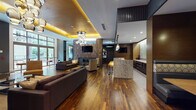 This elegantly designed single-floor amenity space blends contemporary sophistication with warm, upscale materials throughout. Rich hardwood flooring flows continuously across the entire space, anchoring the various functional zones and tying the design together cohesively.

The main lounge area is a standout, centered around a dramatic floor-to-ceiling textured stone accent wall that frames a sleek linear fireplace and wall-mounted flat-screen television. A sculptural cluster chandelier with polished globe fixtures highlights the coffered ceiling, which features warm ambient uplighting in a striking gold tone. Comfortable sectional seating and curated accent chairs complete this inviting gathering zone.

Adjacent to the lounge, a full wet bar commands attention with a substantial granite island, dark cabinetry, an under-counter refrigerator, and a dazzling textured glass backsplash illuminated from behind. Glass pendant lights add to the refined atmosphere.

A quieter alcove with sheer-curtained floor-to-ceiling windows offers a serene retreat with views of lush greenery, furnished with a sleek daybed and ottoman. The space also features a well-lit corridor connecting to additional areas, finished with warm wood paneling and stone tile accents.

Expansive glazed doors open directly onto an outdoor patio with high-top seating, allowing natural light to flood the interior and creating a seamless indoor-outdoor connection.