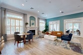 Located at 906 Beacon Square Ct, this single-floor leasing office and amenity center offers a generously sized and thoughtfully designed space for both community residents and prospective guests. The main lounge serves as the centerpiece of the facility, featuring rich teal accent walls, crown molding, and light hardwood flooring throughout. A classic white-mantled fireplace anchors one end of the room, flanked by elegant arched niches and plantation-shuttered windows that flood the space with natural light. Comfortable seating arrangements — including a cognac leather sofa, accent chairs, and a dining table — create a warm, inviting atmosphere. The private office area is bright and functional, with large multi-pane windows overlooking greenery outside, high ceilings, and a spacious conference-style table suited for meetings or leasing consultations. A well-appointed kitchenette hallway features white shaker cabinetry, quartz countertops, stainless steel appliances, and pendant lighting. The fitness center is equipped with treadmills, ellipticals, and a rowing machine, accented by natural light and wall-mounted media. A standout feature is the full indoor racquetball court with a polished hardwood floor and glass entry doors, offering a premium recreational amenity rarely found in a leasing facility.