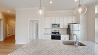 Located in Morehead City, North Carolina, this single-floor residence offers a thoughtfully designed layout with contemporary finishes throughout. The open-concept kitchen is a standout feature, showcasing white shaker cabinetry, a herringbone tile backsplash, stainless steel appliances, and a generous granite island with an undermount sink illuminated by globe pendant lighting. Large windows allow abundant natural light to fill the space, with views of surrounding greenery. Light wood-look flooring runs through the main living areas, complemented by crown molding that adds a refined touch. The living area opens directly to a private balcony through a glass-panel door, further enhancing the airy, open feel. Bedrooms are carpeted for comfort and feature oversized windows with transom panels that maximize daylight. A spacious walk-in closet with wire shelving provides generous storage. The bathroom is well-appointed with a double-sink vanity topped with granite, white shaker cabinetry, wood-look flooring, and a tiled shower-tub combination with subway tile surround and dual mirrors with bold dark frames. The overall palette of warm neutrals and crisp whites gives the space a clean, modern, and welcoming ambiance throughout.