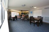 Located at 1 Harbor Ct in Portsmouth, Virginia, this single-floor community space offers a thoughtfully designed layout that accommodates both social gatherings and everyday functional needs. The lounge area is a standout feature, boasting a row of expansive windows that frame captivating views of the neighboring harbor and waterfront. Comfortable seating, coordinated teal accent décor, and warm cove lighting along the crown molding create an inviting and polished atmosphere. Adjacent to the lounge, the dining area continues the refined aesthetic with patterned blue carpeting, recessed cove lighting, and a centrally positioned dining table surrounded by dark wood chairs — ideal for group meals or meetings. The fully equipped kitchen includes warm oak cabinetry, light stone countertops, a full range with overhead microwave, and hardwood-style flooring, providing all the essentials for food preparation. A separate lobby and waiting area features a mix of seating options, including a leather bench and upholstered chairs, alongside indoor greenery that adds a natural touch. This area also connects to a multipurpose room furnished with tables, chairs, and a vending machine, making it well-suited for community use. Throughout the space, consistent lighting, neutral tones, and practical furnishings contribute to a cohesive and functional environment.