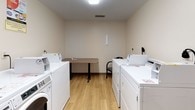 This shared laundry room is a practical, well-organized facility designed to accommodate multiple users efficiently. The space features light hardwood flooring and neutral beige walls that create a clean, bright atmosphere complemented by overhead lighting and a ceiling-mounted smoke detector.

Along the walls, multiple top-loading and front-loading washers and dryers are arranged to maximize capacity, with each machine clearly labeled and equipped with coin or card-operated payment systems. A dedicated utility sink with a faucet is positioned along one wall, providing a convenient hand-washing or pre-treatment station. A laundry rules sign is posted nearby to guide users.

A centrally placed folding table with a laminate surface offers a functional workspace for sorting and folding laundry. Seating is provided in the form of a cushioned chair, allowing users to wait comfortably during wash cycles.

The room is accessed through a glass-paneled door that connects to a hallway, with an adjacent window offering a view into the corridor, enhancing the sense of openness. The overall layout is efficient and straightforward, prioritizing functionality and ease of use for shared occupancy.