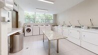Located at 6030 Bathurst Street in Toronto's North York neighbourhood, this shared laundry facility is a clean, efficiently organized space designed to serve building residents with convenience and comfort. The room is lined with a generous arrangement of commercial-grade top-load and front-load washing machines, complemented by a full row of dryers, providing ample capacity for simultaneous use. A large granite-topped folding table anchors the centre of the room, offering a practical surface for sorting and folding laundry. The space is finished with light-toned large-format porcelain tile flooring, contributing to a bright and hygienic atmosphere. Natural light streams in through a wide bank of windows along one wall, offering pleasant views of the surrounding greenery and keeping the space feeling open and airy. Overhead fluorescent lighting ensures the room remains well-lit at all hours. A utility sink is also present, adding further functionality. Coin-operated or card-based payment terminals are mounted on the machines for easy access. Informational notices and a waste receptacle are thoughtfully positioned near the entry door, maintaining an organized and user-friendly environment. Overall, this laundry room is a well-maintained, practical amenity that reflects a high standard of building management.