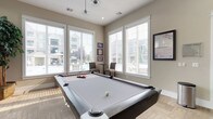 This single-level game room is designed as a dedicated recreational space, anchored by a full-size pool table with a sleek grey felt surface and dark wood frame. Large, white-trimmed windows line multiple walls, flooding the room with natural light and offering views of the surrounding outdoor pool area and landscaping. White horizontal blinds provide adjustable light control throughout the day.

The flooring features a neutral, tile-patterned surface that complements the room's warm beige and taupe color palette. A pendant light fixture hangs above the pool table, providing focused overhead illumination. One wall showcases a stacked stone accent, against which a large flat-screen television is mounted, making the space equally suited for watching sports or socializing. A display shelf above the TV holds decorative items and greenery, adding character to the room.

Seating is thoughtfully incorporated, with tufted upholstered bar-height chairs and small bistro-style tables positioned near the windows, creating comfortable spots to relax between games. Framed artwork and sports memorabilia decorate the walls, contributing to the room's casual and lively atmosphere. The overall design balances functionality with a welcoming, community-oriented ambiance.