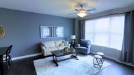 This single-story space centers around a thoughtfully designed living room that combines comfort and contemporary style. The room is distinguished by its cool blue-grey walls, which create a calm and cohesive atmosphere throughout. Dark hardwood-style flooring grounds the space and contrasts nicely with the light-toned furnishings, including a cream sofa adorned with decorative pillows and a coordinating blue accent chair. A rectangular dark wood coffee table anchors the seating area, which is further defined by a large patterned area rug in soft blue and white tones. Natural light filters generously into the room through large windows dressed with horizontal blinds and floor-length drapes, lending an airy quality to the interior. A ceiling fan with a built-in light fixture provides both practical ventilation and ambient lighting. Two matching framed artworks on the main wall add a polished, gallery-like touch. Accent pieces, including metallic side tables and decorative ottomans, contribute to the layered, curated feel of the space. Overall, the living area presents a well-balanced blend of functionality and refined design.