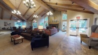 Located at SW Barnes Rd in West Haven, Oregon, this inviting single-floor clubhouse is defined by its striking architectural character and warm, lodge-inspired aesthetic. The centerpiece of the main lounge is a soaring vaulted ceiling with exposed timber beams and white-painted wood paneling, illuminated by elegant chandelier lighting and recessed track fixtures. Large windows throughout flood the interior with natural light and frame lush, tree-lined views of the surrounding Pacific Northwest landscape.

The main gathering area features a dramatic stone fireplace flanked by tall windows, creating a cozy focal point surrounded by ample seating arrangements. Stone tile flooring flows throughout, grounding the space with a natural, durable finish. A dedicated dining area offers multiple round tables with tufted leather chairs, complemented by a built-in window bench seat that overlooks the verdant outdoors — ideal for casual gatherings or meetings.

A quiet reading nook with wingback chairs and a bookshelf provides a more intimate retreat, bathed in natural light from a row of windows. The space also includes a kitchenette with warm wood cabinetry, a stone countertop bar with seating, pendant lighting, and built-in appliances, making it well-equipped for events and social functions. Glimpses of an outdoor pool area suggest additional amenity access just beyond the interior.