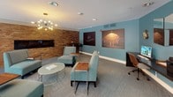 Located at 10001 E Dry Creek Rd in Englewood, Colorado, this thoughtfully designed single-floor community lounge blends contemporary style with comfortable functionality. The main seating area is anchored by plush teal upholstered chairs and sofas arranged around sleek glass and wood accent tables, all beneath a striking sputnik chandelier that serves as a visual centerpiece. A textured brick feature wall adds warmth and character, complemented by large-format music-themed artwork that lends the space a creative, artistic ambiance.

A built-in fireplace with a wall-mounted television is seamlessly integrated into the brick accent wall, creating an inviting focal point ideal for relaxed gatherings. Patterned carpet flooring ties the space together throughout.

Floor-to-ceiling sliding glass doors along one wall flood the interior with natural light and provide direct access to an outdoor pool area, blurring the boundary between indoor comfort and outdoor leisure. A casual dining nook near these doors features a round wood table with designer chairs, perfect for smaller gatherings.

A dedicated workstation area, accented by a teal feature wall and jazz-inspired neon artwork, offers a desktop computer and ergonomic seating — a practical space for residents needing a quiet place to focus. Overall, the lounge presents a vibrant, music-inspired aesthetic with versatile spaces for socializing, dining, and working.