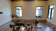 This single-floor fitness space is purpose-built for indoor cycling workouts, offering a clean and focused environment for exercise. The room features rich hardwood-style flooring that adds warmth and durability underfoot, well-suited to the demands of an active fitness setting. Multiple stationary cycling bikes are arranged throughout the space, providing ample room for individual workouts or group cycling sessions. Two large windows with dark trim frames are positioned symmetrically along the wall, allowing natural light to fill the room and creating an open, energizing atmosphere. The neutral-toned walls complement the overall aesthetic, keeping the environment visually calm and distraction-free. The ceiling features warm-toned wood detailing that adds an architectural accent and a sense of refinement to the space. The layout is straightforward and functional, prioritizing ease of movement and accessibility around the equipment. Overall, this fitness room is a well-appointed, light-filled space designed to support a focused and motivating cycling workout experience.