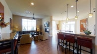 This single-floor space is designed as an open-concept gathering area that seamlessly blends multiple functional zones into one cohesive layout. The living lounge area anchors one side of the space, featuring a wall-mounted flat-screen television above a sleek fireplace, complemented by a ceiling fan and an abundance of natural light streaming through large windows. Comfortable seating, including a dark sectional sofa and neutral accent chairs, is arranged around a patterned area rug atop rich, warm hardwood flooring that extends throughout the entire space.

Transitioning toward the opposite end, a bar counter with leather-upholstered barstools serves as a social hub, illuminated by stylish pendant lights hanging above the granite countertop. Beyond the bar, a billiards table takes center stage beneath additional pendant lighting, with natural light pouring in through glass French doors that provide views of the outdoors. The warm, neutral wall tones and recessed lighting throughout create a welcoming and versatile ambiance, making this space well-suited for both casual relaxation and social gatherings.