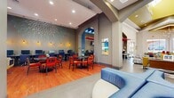 This single-story clubhouse is designed with a bold, contemporary aesthetic that combines vivid color accents with modern architectural details throughout its multiple functional zones. The main lounge area features soaring ceilings and expansive floor-to-ceiling windows that flood the space with natural light and offer open views of the surrounding landscape. A large sectional sofa in deep blue, accented with red and olive throw pillows, anchors the seating area alongside a flat-screen display and a sleek fireplace feature wall finished in white textured tile with a gold tray ceiling above. Adjacent to the lounge, a dining and social area offers hardwood-style flooring, round tables, and vibrant red chairs beneath a striking geometric wall mural rendered in teal and white. This area flows into a well-equipped business center with multiple desktop workstations, warm amber pendant lighting, and wood flooring. An arched passageway connects the social zones seamlessly. The fitness center is a standout feature, boasting bold red and yellow upper walls, rows of cardio machines including ellipticals and treadmills, strength and resistance equipment, and large windows providing natural light and exterior views. Patterned carpet tiles complete the gym's energetic atmosphere.