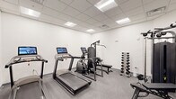 Located at 250 Celebration Ct in Middletown, Delaware, this fitness center offers a well-organized exercise room designed to support a comprehensive workout routine. The space features a clean, modern aesthetic with crisp white walls, a drop-panel ceiling with recessed fluorescent lighting, and durable speckled rubber flooring suited for high-traffic athletic use. A large wall-mounted mirror expands the visual depth of the room while allowing users to monitor their form during exercise. The equipment selection is thoughtfully curated, including multiple treadmills equipped with digital touchscreen displays, an elliptical machine, a multi-station cable and strength training system with an adjustable bench, a rowing machine, and a rack of free-weight dumbbells spanning a range of sizes. Entry to the room is through a sleek black-framed glass door, which provides visibility into the adjacent corridor and contributes to the space's modern, professional appearance. The layout ensures clear circulation between equipment stations, making it practical for individual use. Overall, the fitness center presents a functional and well-maintained environment that meets the essential needs of residents or occupants seeking convenient access to both cardio and strength training equipment.