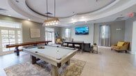 Located at 10880 Angel Fish Way in Jacksonville, Florida, this single-floor community clubhouse offers a well-appointed gathering space designed for both recreation and relaxation. The centerpiece of the space is a striking game room anchored by a full-size pool table beneath a modern pendant light fixture and a shuffleboard table, all set atop a decorative area rug over large-format light tile flooring. A dramatic tiered tray ceiling with recessed lighting crowns the game area, adding architectural depth and elegance to the room. Natural light flows generously through multiple windows dressed with sheer and patterned curtains, enhancing the bright, airy atmosphere. A comfortable lounge area features a sectional sofa, accent chairs in warm mustard tones, and a wall-mounted flat-screen television, creating an inviting space for casual gatherings. A dedicated refreshment station includes white shaker cabinetry, granite-style countertops, a stainless utility sink, open shelving, a beverage refrigerator, and a commercial-grade coffee machine, making it ideal for entertaining. Soft gray walls, curated coastal artwork, and tasteful decorative accents throughout lend the space a polished yet welcoming character. French doors provide access to the exterior, further connecting the interior to its surroundings.