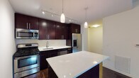 This single-floor unit showcases a thoughtfully designed layout with consistent dark hardwood flooring throughout the main living areas, creating a cohesive and polished aesthetic. The kitchen serves as a standout feature, equipped with rich dark-stained cabinetry extending to the ceiling for ample storage, a crisp white quartz island, white tile backsplash, and a full suite of stainless steel appliances including a gas range, built-in microwave, dishwasher, and a French door refrigerator with a water and ice dispenser. Pendant lights and a track lighting system illuminate the space effectively. A wide hallway connects the living areas to the private quarters, which include a generously sized bedroom with two large closets offering built-in shelving and rod storage. A floor-to-ceiling glass door in the living area floods the space with natural light and provides outdoor access with urban views. Two bathrooms are finished with granite-topped dark vanities, tile flooring, and large mirrors. The full bath features a walk-in glass-enclosed shower with subway-style tile walls. Clean white walls and a neutral palette throughout contribute to a bright, modern ambiance.