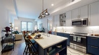 This thoughtfully designed single-floor residence blends modern aesthetics with functional living. The heart of the home is a striking kitchen outfitted with two-tone cabinetry — bold navy lowers paired with soft light gray uppers — accented by brushed gold hardware throughout. A generous island with a quartz waterfall countertop serves as both a prep space and casual dining area, illuminated by an industrial-style trio of pendant lights. A herringbone tile backsplash, under-cabinet lighting, a gold faucet, and a full suite of stainless steel appliances, including a French door refrigerator, complete the culinary space. The kitchen flows seamlessly into an open living area bathed in natural light from large windows and a glass-panel door leading to a private balcony. The bedroom offers a serene retreat with plush carpeting, a bright window, and generous wall space. The bathroom features neutral gray tile, a tub-shower combination, a white vanity with quartz countertop, and gold accents that echo the home's cohesive design language. Light wood-style flooring runs throughout the main living areas, tying together a space that balances contemporary style with everyday comfort.