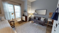 This single-floor studio offers a well-considered open-concept layout that seamlessly integrates the living, sleeping, and kitchen areas into one cohesive space. The living area is anchored by warm hardwood-style flooring, a plush textured area rug, and a contemporary gray sofa accented with decorative pillows. A sleek acrylic coffee table and a modern rocking chair contribute to the eclectic yet functional aesthetic, while a large window dressed with sheer curtains invites abundant natural light. The accent wall in a muted blue-gray tone adds depth and visual interest, complemented by a wall-mounted mirror that enhances the sense of openness. A mid-century style dresser and decorative shelving complete the living zone with practical storage. The kitchen is efficiently appointed with stainless steel appliances, including a range and over-the-range microwave, dark granite-style countertops, and crisp white cabinetry that maximizes storage. The sleeping area flows naturally from the kitchen, featuring a full-sized bed positioned beneath a window that fills the space with soft, natural light. A neutral color palette, clean lines, and a cohesive mix of modern and mid-century furnishings give this studio a warm, inviting ambiance throughout.