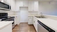 This single-floor space is designed with a clean, functional layout that emphasizes an open and cohesive feel throughout. Wood-look flooring runs continuously across the main living areas, offering a warm, unified aesthetic that ties the space together.

The living area is generously proportioned, featuring neutral white walls, clean baseboards, and a ceiling fan that contributes to the room's comfort. A sliding glass door leads to an outdoor area, allowing natural light to filter in and providing a connection to the exterior.

The kitchen is positioned in an open-concept arrangement, partially separated from the living area by a breakfast bar counter that offers casual seating. White shaker-style cabinetry extends from floor to ceiling, providing ample storage, while light-toned countertops keep the space feeling bright and clean. Stainless steel appliances, including a range and microwave, are complemented by a double basin sink and a dishwasher.

A hallway connects the main living spaces to the remaining rooms, with a door visible that opens to additional areas. The bathroom includes a tub-shower combination finished in crisp white tile surround, maintaining the space's clean, minimal aesthetic. Throughout, the design prioritizes practicality and a light, airy atmosphere.
