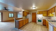 This single-floor home offers a functional and well-organized layout designed for comfortable everyday living. The kitchen serves as a central hub of the space, featuring warm honey-toned oak cabinetry that extends both above and below the countertops, providing generous storage throughout. A peninsula with a breakfast bar connects the kitchen to the living area, creating an open-concept flow. White appliances, including a freestanding electric range, dishwasher, and refrigerator, are all included. Tile flooring runs throughout the kitchen, while the remainder of the home is finished with wall-to-wall carpet in a neutral tone. The spacious living room benefits from abundant natural light, courtesy of a sliding glass door that opens to an outdoor deck or patio, as well as a secondary window framing views of the surrounding greenery. A ceiling fan with a light kit provides both comfort and illumination. The bedrooms are well-proportioned, carpeted, and finished with warm wood-trimmed windows and doors that complement the home's consistent aesthetic. The bathroom features a clean, full-size tub and shower combination. A chandelier in the dining area adds a touch of warmth, completing this practical and inviting single-level home.
