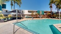 This residential complex in Los Alamitos, California, showcases a blend of modern architecture and resort-style amenities. The exterior is characterized by a striking combination of white and teal-colored facades, creating a visually appealing contrast. The complex features a large, inviting swimming pool as its centerpiece, surrounded by a spacious concrete deck area ideal for sunbathing and relaxation.

The landscaping incorporates mature palm trees, adding a tropical ambiance to the property. Security is evident with a gated entrance and perimeter fencing. The building's design includes interesting architectural elements such as protruding window bays, covered balconies, and varying rooflines, adding visual interest and dimension to the structure.

Ample outdoor seating, including lounge chairs and tables, is provided around the pool area. The complex appears to be multi-storied, offering residents elevated views. The overall atmosphere suggests a focus on outdoor living and community spaces, making it an attractive option for those seeking a blend of comfort and resort-style living in a suburban setting.