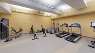 This fitness center offers a comprehensive workout environment thoughtfully organized across a single floor. The space is divided into distinct functional zones, each catering to a different style of training. The cardio area is lined with treadmills, elliptical trainers, rowing machines, and a stationary bike, providing ample options for aerobic exercise. A ceiling fan overhead contributes to comfortable airflow throughout this section.

The strength and functional training zone features a large multi-tier power rack stocked with kettlebells, medicine balls, weight plates, and battle ropes, alongside a dedicated dumbbell rack with a full range of free weights and an adjustable workout bench. A wall-mounted mirror expands the sense of space and allows for form monitoring during exercises. Additional functional training accessories, including stability balls and foam rollers organized on a vertical storage rack, round out the offerings.

The flooring throughout is durable speckled rubber, well-suited for high-traffic fitness use. Overhead fluorescent and recessed lighting ensures the entire space is brightly and evenly illuminated. The warm yellow-toned walls contribute to an energizing yet welcoming atmosphere. The layout is open and navigable, with clear sightlines across the room, making the fitness center both practical and easy to use.