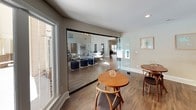 This single-story space is thoughtfully designed to serve as a communal gathering and business hub, combining multiple functional areas within an open, cohesive layout. Wood-look flooring flows continuously throughout, unifying each zone and contributing to a warm, inviting ambiance.

The main lounge area is bright and airy, benefiting from large windows and glass-paneled French doors that invite abundant natural light and provide views of an outdoor pool area. Mid-century modern-inspired seating, including accent chairs and round bistro tables, is arranged to encourage both casual socializing and quiet relaxation. A bold navy blue accent wall anchors the seating area, adding a strong design focal point against the neutral taupe walls.

A glass partition wall with a sleek black-framed sliding door separates a secondary lounge or meeting area from the main space, maintaining visual openness while offering a degree of privacy. Framed artwork with teal accents adorns the walls, complementing the overall color palette.

The dedicated business center features a long granite-topped workstation built into rich wood cabinetry, accommodating multiple workstations with ergonomic chairs and computer monitors. Recessed lighting and a blue accent wall give this area a focused, professional atmosphere, making it well-suited for productivity.