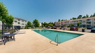 This outdoor community pool area in Augusta, Georgia offers a well-appointed recreational space designed for relaxation and leisure. At its center sits a generously sized rectangular swimming pool with clear, blue-green water, accessible via stainless steel handrails and marked with depth indicators for safety. The surrounding concrete sun deck provides ample space for lounge chairs, allowing residents to enjoy the Georgia sunshine in comfort.

The pool area is fully enclosed by a black wrought iron fence, providing both security and a clean aesthetic boundary. Gated entry points are clearly marked, and safety signage is posted at the entrance. A pergola structure near the pool area offers a shaded retreat, complemented by outdoor patio furniture including a table and chairs — ideal for gathering and socializing.

Well-maintained green lawns border the fenced perimeter, dotted with mature trees that provide natural shade and a pleasant visual backdrop. The surrounding multi-story residential buildings are visible from within the pool area, reinforcing the community-oriented nature of the space. The open-air setting benefits from abundant natural light and broad views of the sky, creating a bright and inviting atmosphere throughout the day.