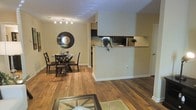 This single-floor residence showcases a well-connected open layout that seamlessly integrates the living, dining, and kitchen areas into one cohesive space. Warm-toned hardwood flooring extends throughout, adding a sense of continuity and a natural aesthetic that anchors the interior.

The living area is defined by a neutral, light-colored area rug and a glass-top coffee table, creating a comfortable and inviting gathering space. Adjacent to this is the dining area, which features a round glass dining table accompanied by dark wood chairs. A large circular mirror with a dark wood frame serves as a striking decorative focal point on the dining room wall, while a track lighting fixture overhead provides targeted illumination.

A half-wall partition with a pass-through opening connects the dining and living areas to the kitchen, allowing light and visibility to flow between the spaces while maintaining a degree of separation. The kitchen, visible beyond the partition, features dark cabinetry and includes a touch of greenery through potted plants, adding a fresh element to the interior. White walls and neutral tones throughout contribute to a bright, clean, and functional atmosphere.