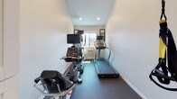 Located at 4179 Third Ave in San Diego's vibrant North Park/Hillcrest neighborhood, this single-floor fitness center offers a well-organized and functional workout space. The room is finished with neutral white walls, clean white baseboards, and durable dark carpet flooring designed to withstand high-traffic athletic use. Recessed lighting throughout provides consistent, even illumination across the entire space. The cardio area is equipped with multiple machines, including a treadmill and an elliptical trainer, each outfitted with individual monitor screens for an enhanced workout experience. A window at the far end of the cardio zone fills the space with natural light and offers an open view of the surrounding San Diego streetscape, creating an airy and motivating atmosphere. On the opposite end of the room, a dedicated strength and functional training area features a weight bench, a rack of medicine balls in varying sizes, and additional equipment organized along a mirrored glass partition. The mirror serves to visually expand the space while allowing users to monitor their form during exercise. The layout is efficient, making thoughtful use of the available floor area to accommodate a variety of workout routines. Overall, the fitness center presents a clean, modern aesthetic with practical amenities suited for daily use.