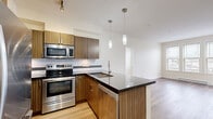 This 2-bedroom, 1-bathroom single-floor unit offers a thoughtfully designed layout that maximizes functionality and comfort. The open-concept kitchen is a standout feature, showcasing warm wood-grain cabinetry, dark granite countertops, a stylish peninsula with an undermount sink, and a full suite of stainless steel appliances including a range, microwave, dishwasher, and refrigerator. Pendant lighting above the peninsula and a tile backsplash with a decorative mosaic accent strip add refined detail to the space. Light wood-tone flooring flows throughout the main living areas, creating a cohesive and contemporary aesthetic. The living area is bright and open, benefiting from natural light and a clean, neutral palette. The primary bedroom is a well-proportioned space featuring large windows with outdoor views and access to an adjacent room. The second bedroom is carpeted, with windows and a baseboard heater, offering a cozy and practical space. The bathroom is finished with large-format tile flooring, a wood-toned vanity with a stone countertop, and a full mirror with overhead lighting. A stacked washer and dryer unit is conveniently located within the unit. A dedicated closet completes this efficiently arranged home.