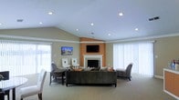 This single-floor space features an open and airy living area defined by its vaulted ceilings and recessed lighting, which together create a bright, welcoming atmosphere. The room centers around a classic white-mantled fireplace set against a warm terracotta-toned accent wall, above which a flat-screen television is mounted, serving as a natural focal point. A second television is also wall-mounted on the opposite side of the room, offering additional viewing options. Large windows dressed with vertical blinds line multiple walls, allowing generous amounts of natural light to fill the space while maintaining privacy. The flooring throughout is carpeted in a neutral tone, contributing to a comfortable and cohesive aesthetic. Seating is arranged thoughtfully, with a substantial sectional sofa positioned to face the fireplace, complemented by upholstered accent chairs and a small side table that provide additional gathering areas. The neutral color palette of beige and gray tones throughout the walls, furnishings, and flooring lends the space a clean, understated elegance. Overall, the layout is well-suited for communal use, offering both open flow and defined seating zones.