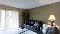 This single-floor space offers a welcoming and thoughtfully designed interior that blends comfort with modern style. The living area serves as the focal point, featuring a sleek black leather sofa accented with patterned throw pillows and complemented by a glass-top coffee table that adds an open, airy feel to the room. The walls are finished in a combination of warm neutral tones and a rich olive green accent wall, creating a grounding, earthy ambiance throughout. Decorative wall-mounted floral accents add a subtle artistic touch to the space. A large window dressed with vertical blinds floods the room with natural light while providing privacy and light control. A wall-mounted air conditioning unit ensures year-round comfort. Lush indoor greenery, including a tall leafy plant in the corner, brings life and freshness to the interior. The flooring features a light-toned area rug over what appears to be a hard surface floor, adding warmth and texture underfoot. The textured ceiling adds a classic finish to the overall design. This space presents a functional and inviting environment suited for relaxed everyday living.