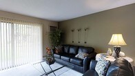 This single-floor space offers a welcoming and thoughtfully designed interior that blends comfort with modern style. The living area serves as the focal point, featuring a sleek black leather sofa accented with patterned throw pillows and complemented by a glass-top coffee table that adds an open, airy feel to the room. The walls are finished in a combination of warm neutral tones and a rich olive green accent wall, creating a grounding, earthy ambiance throughout. Decorative wall-mounted floral accents add a subtle artistic touch to the space. A large window dressed with vertical blinds floods the room with natural light while providing privacy and light control. A wall-mounted air conditioning unit ensures year-round comfort. Lush indoor greenery, including a tall leafy plant in the corner, brings life and freshness to the interior. The flooring features a light-toned area rug over what appears to be a hard surface floor, adding warmth and texture underfoot. The textured ceiling adds a classic finish to the overall design. This space presents a functional and inviting environment suited for relaxed everyday living.