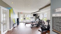 This fitness center is designed to provide a functional and inviting workout environment. The space is finished with rich, dark hardwood-style flooring throughout, lending a polished aesthetic that contrasts cleanly with the light gray and white walls. A textured ceiling with recessed lighting ensures the room is well-lit, while multiple windows along the perimeter allow abundant natural light and outward views of the surrounding greenery.

The main workout area houses a selection of BodyCraft cardio equipment, including a treadmill, elliptical trainer, rowing machine, and a recumbent stationary bike. A wall-mounted flat-screen television is positioned near the cardio machines for entertainment during workouts. A secondary area provides space for strength training, featuring an adjustable workout bench and a full-length mirror, making it suitable for a variety of exercise routines.

A bold geometric accent wall in shades of green, blue, and gray adds a modern, energetic visual element near the entrance. The space also includes practical amenities such as sanitizing dispensers, a waste bin, and clearly marked emergency exits, ensuring both convenience and safety for users.