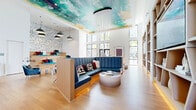 Located at 2525 Paseo Park Rd in Orlando, Florida, this single-floor communal space is a striking example of contemporary interior design, blending artistic expression with functional amenities. The layout is generously proportioned, offering multiple distinct seating and activity zones that flow seamlessly across light-toned hardwood flooring throughout.

The centerpiece of the main lounge area is a bold, hand-painted ceiling mural awash in teal, gold, and deep blues, anchored by a dramatic chandelier composed of cascading white strands. A large L-shaped banquette upholstered in rich navy blue provides a prominent gathering spot, complemented by a sculptural round ottoman. Built-in shelving units line one wall, displaying curated décor alongside a mounted flat-screen television.

An amphitheater-style stepped seating area, adorned with colorful cushions and backed by an abstract line-art wallcovering, offers a casual space for group presentations or relaxed socializing. Oversized pendant lights hang above this zone, adding a warm, artistic touch.

A separate café-style area features high-top bistro tables with leather barstools set against a teal brick accent wall. The kitchen nook is equipped with cabinetry, countertops, appliances, a beverage station, and a wall-mounted television. Floor-to-ceiling windows throughout invite abundant natural light and views of the surrounding outdoor environment.
