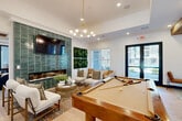 Located in Glendale, Arizona, this single-floor community amenity space seamlessly combines a living lounge and a fully equipped kitchen into one cohesive, design-forward environment. The living area is anchored by a striking floor-to-ceiling green tile accent wall, which frames a linear electric fireplace and a wall-mounted television. Comfortable neutral-toned seating surrounds a glass-top coffee table, creating an inviting gathering spot, while a full-size billiard table adds a recreational focal point to the room. A large vertical garden panel and natural light from oversized windows contribute to a fresh, lively atmosphere, with a glass door providing direct views to the exterior. The kitchen is equally impressive, featuring exposed wood ceiling beams that lend warmth and texture overhead. Dark cabinetry with gold hardware contrasts elegantly against a white hexagonal tile backsplash and bright white countertops. A spacious central island with bar-height rattan seating serves as a social hub, while open shelving, a coffee station, beverage refrigerator, and built-in display shelving with bold navy tile accents complete the space. Bold geometric floor tile in the kitchen transitions to light wood-look flooring throughout the lounge, tying both areas together with a modern yet welcoming aesthetic.