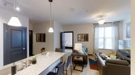 This single-floor residence offers a thoughtfully designed open-concept layout that seamlessly connects the main living areas. The kitchen features a generous granite-topped island with bar seating, illuminated by a stylish pendant light, making it an ideal space for casual dining and entertaining. Sleek cabinetry and modern finishes complete the culinary space.

The living area flows naturally from the kitchen, creating an inviting and cohesive gathering space. A ceiling fan overhead provides comfort, while large windows dressed with sheer curtains allow generous natural light to fill the room. The space is furnished with comfortable seating and accented by a colorful area rug, adding warmth and personality to the interior.

A bedroom is visible directly off the main living area, offering convenient access and a sense of openness within the floor plan. Throughout the residence, neutral wall tones create a calm, versatile backdrop that complements a variety of décor styles.

The overall floor plan, as seen from above, reveals a well-organized layout with clearly defined zones for living, sleeping, and utility — all connected by a logical and accessible flow. The combination of modern design elements, ample natural light, and a functional open-concept arrangement makes this a comfortable and stylish single-floor living space.