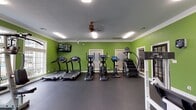 This fitness center offers a comprehensive workout environment spread across a single, open floor. The space is immediately distinguished by its bold green walls, white crown molding, and a textured ceiling, creating an energetic yet structured atmosphere. Rubber flooring throughout provides both durability and safety underfoot.

The cardio zone is well-stocked with multiple treadmills, elliptical trainers, a stair climber, and a stationary bike, each equipped with individual digital consoles for personalized workout tracking. A wall-mounted flat-screen television accompanies the cardio area, adding an entertainment option during exercise sessions.

The strength training section features a multi-station cable machine, a dedicated leg press and leg extension machine, and an additional selectorized strength unit. A full rack of dumbbells and various free weight equipment, including barbells and weight plates, are also present. A large wall mirror spans one side of the room, enhancing the sense of space and allowing users to monitor their form.

Natural light enters through several windows, offering views to the surrounding exterior. A ceiling fan provides additional air circulation, complementing the overhead fluorescent lighting. The overall layout is efficiently organized, maximizing floor space and accommodating a variety of fitness routines simultaneously.