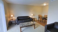 This single-floor residence offers a practical and welcoming layout designed for everyday comfort. The living and dining areas flow seamlessly together, creating an open and functional shared space. The living area features plush carpeting underfoot and is furnished with a sleek black leather sofa, a stone-top coffee table, and side tables with warm lamp lighting that contribute to a cozy ambiance. Neutral-toned walls keep the space feeling bright and airy, while the flush ceiling light provides even illumination throughout. Transitioning toward the dining area, the warm wood-style flooring adds a touch of character and visual contrast to the soft carpeted zone. A glass-top dining table surrounded by classic wooden chairs provides a clean and inviting space for meals. The overall design aesthetic is straightforward and understated, with a neutral color palette and modest furnishings that lend the space a calm, livable quality. The layout is efficient, making good use of the available floor space while maintaining an uncluttered feel throughout.