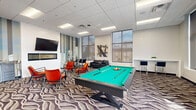Located at 295 Polifly Rd in Hackensack, NJ, this well-appointed community lounge offers a generously sized, single-floor open layout designed for socializing and recreation. The space is anchored by a striking patterned carpet with flowing wave motifs that extends throughout, lending a dynamic visual energy underfoot. Bold orange upholstered lounge chairs paired with glass-top coffee tables create an inviting seating area, complemented by a sleek black leather sofa. A full-size pool table with a vibrant green felt surface serves as a central recreational feature. One wall showcases an eye-catching cityscape mural with vertical stripe accents, above which a wall-mounted flat-screen TV is installed, adding an entertainment focal point. Large floor-to-ceiling windows on multiple walls flood the room with natural light and provide open outdoor views. A compact kitchenette along one wall features dark cabinetry and countertop appliances, offering convenient refreshment access. The entrance is framed by modern glass-panel double doors, contributing to the space's contemporary aesthetic. Recessed panel ceiling tiles with integrated lighting complete the clean, modern design, making this lounge a versatile and inviting gathering space.