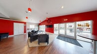 The Villas clubhouse at 1904 S Rouse St in Pittsburg, Kansas, serves as the central amenity hub for the community. The single-floor facility is thoughtfully laid out to accommodate a variety of resident needs. The heart of the space is a spacious communal lounge featuring vaulted ceilings, warm hardwood-style flooring, and bold red accent walls that give the room an energetic character. Vibrant red pendant lighting hangs from the ceiling, complementing the lively aesthetic. A signature hand-painted mural — depicting ocean waves, palm trees, and the community's "The Villas" branding — serves as a standout focal point, adding a fun and personalized touch. The lounge is furnished with comfortable dark sofas and chairs arranged around a central coffee table, alongside high-top bar seating. Dual flat-screen TVs are mounted for entertainment. Large glass doors flood the space with natural light and provide direct access to an outdoor pool and patio area. Additional amenities include a dedicated tanning room with a full tanning bed set against vibrant orange walls, a well-equipped communal laundry room lined with rows of washers and dryers under bright overhead lighting, and a mailroom corridor featuring an extensive bank of resident mailboxes. A hallway connects the various rooms, ensuring smooth flow throughout the facility.