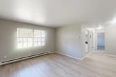 Located at 416 Lawrence Ave in North Syracuse, NY, this one-bedroom, one-bathroom unit offers a thoughtfully arranged single-floor layout with a clean, modern aesthetic throughout. The living room is a bright, open space finished with light-toned luxury vinyl plank flooring and neutral gray walls, creating a welcoming atmosphere. Large windows along the exterior wall invite generous natural light and offer views of the surrounding greenery. A baseboard heating system runs along the perimeter, ensuring year-round comfort. The layout flows naturally from the living area toward the kitchen, which is visible in the background with white cabinetry. The spacious bedroom features soft wall-to-wall carpeting, a window-mounted air conditioning unit, and a double-door closet, providing ample storage. Natural light enters through a multi-pane window, adding to the room's airy feel. The bathroom is finished to a modern standard, showcasing a white vanity with a framed mirror, matte black fixtures, and a full bathtub with a bright window above. Light wood-style flooring ties the space together cleanly. Overall, this unit presents a well-maintained, move-in-ready living space with practical features and a cohesive, neutral design palette throughout.