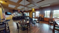 Located at 4701 Seventh Ave SW in Olympia, Washington, this pub offers a comfortable and inviting atmosphere across a single, well-appointed floor. The space is anchored by a curved, dark-wood bar with ample seating along its perimeter, backed by a mirrored display and pendant lighting that highlights the full service area. A wall-mounted flat-screen television is positioned nearby for easy viewing.

The dining area features rich hardwood flooring throughout and a thoughtfully designed coffered ceiling with recessed panels and warm accent lighting, adding architectural character to the room. Seating options are varied, including standard tables with ladder-back chairs, high-top bar tables with stools, and cushioned booth-style banquette seating along one wall — accommodating both casual drop-ins and longer gatherings.

Large windows line the walls, flooding the space with natural light and offering views of the surrounding neighborhood. A set of double glass doors provides additional access and further enhances the connection to the outdoors. Decorative touches include vintage-style framed posters, colorful banners, and striped upholstery on the booth seating, contributing to the pub's eclectic and relaxed character. Track lighting supplements the pendant fixtures, ensuring the space is well-lit throughout the day and evening.