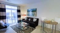 This single-floor space presents a thoughtfully designed living area that balances comfort and contemporary style. The living room serves as the focal point, anchored by a dark upholstered sectional sofa accented with patterned throw pillows. A glass-top coffee table sits atop a bold circular-print area rug, adding visual interest to the carpeted floor. A vibrant abstract canvas depicting a reflective forest scene adorns the main wall, providing a striking pop of color against the neutral white walls. Natural light flows generously into the space through a large window dressed with bold black-and-white striped curtains, which opens to an adjoining balcony, offering a seamless connection to the outdoors. The dining area is integrated directly into the living space, featuring a high bar-style table with modern stools — an efficient use of the available footprint. The overall ambiance is modern and inviting, with a neutral base palette enlivened by teal and blue accents throughout the décor. The layout is open and practical, making efficient use of the space while maintaining a clean, well-organized feel.