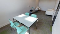 The Lounge is a thoughtfully designed single-floor space that combines modern aesthetics with functional versatility. The interior is characterized by bright white walls and a patterned carpet featuring a neutral grey base accented with vibrant green geometric detailing, adding a lively visual element to the overall clean design.

The space is divided into two distinct functional zones that flow seamlessly into one another. A collaborative work area features a sleek white rectangular table with chrome legs, surrounded by modern mint-green chairs — ideal for group meetings or focused work sessions. Natural light enters through windows at the far end of the room, supplemented by recessed ceiling lights and flush skylight panels that keep the space well-illuminated throughout.

The lounge area is furnished with a tufted white leather sofa and a pair of sculptural swan-style swivel chairs in white with polished chrome bases, creating an inviting setting for casual conversation or relaxed seating. A slim black utility cart with a teal desk lamp and small accessories adds a practical touch to the seating nook.

Overall, The Lounge maintains a crisp, contemporary ambiance with a cohesive color palette of white, grey, and mint green, making it a welcoming and adaptable space suited for both productive work and informal social interaction.