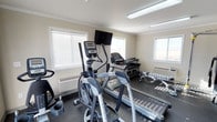 This fitness center is a compact yet fully functional exercise space designed to support a well-rounded workout routine. The room is finished with light-colored walls accented by crown molding, and the speckled rubberized flooring provides both durability and safety underfoot. Overhead fluorescent lighting combines with multiple windows dressed in horizontal blinds to create a bright and energizing atmosphere throughout the space.

The cardio area is well-appointed, featuring Matrix-brand elliptical trainers, at least one treadmill, and a stationary bike, offering a variety of options for aerobic exercise. A wall-mounted flat-screen television is positioned prominently, allowing users to stay entertained during workouts. Toward the back of the room, a dumbbell rack holds a full range of free weights alongside a weight bench, catering to strength training needs.

Climate comfort is addressed with a wall-mounted HVAC unit, ensuring the space remains at a comfortable temperature year-round. The single-floor layout keeps everything accessible and easy to navigate. Overall, this fitness center strikes a balance between practicality and functionality, making it a convenient and well-outfitted facility for daily exercise.