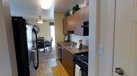 This single-floor residence offers a well-designed layout that balances practicality and comfort. The kitchen is a standout feature, showcasing rich warm-toned wood cabinetry that extends to the upper reaches of the walls, providing generous storage space. Dark stone-look countertops complement the cabinetry, while black appliances — including a gas range and dishwasher — lend a cohesive, modern aesthetic. A stainless steel sink sits neatly beneath upper cabinets, and a skylight overhead floods the kitchen with natural light. Decorative greenery atop the upper cabinets adds a lively touch to the space. The kitchen flows seamlessly into the dining area, which is anchored by a classic dining table and chairs set on a area rug. A ceiling fan with a light fixture keeps the dining space comfortable, while a large window ushers in abundant natural light, creating an airy and inviting atmosphere. Tile flooring in the kitchen transitions naturally into the adjoining spaces, tying the home's design together. Overall, this residence offers a warm, functional environment with thoughtful design details throughout.