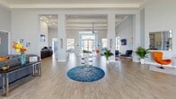 This single-floor community clubhouse offers a bright, open-concept design characterized by soaring ceilings, wide-plank tile flooring, and an airy, welcoming ambiance throughout. The grand entrance features floor-to-ceiling glass doors and transom windows that flood the foyer with natural light, framing views of the surrounding outdoor landscape. A custom branded entryway rug greets visitors as they step inside.

The central atrium-style common area serves as the heart of the space, anchored by a sculptural glass-top table with a chrome base set atop a bold circular blue rug. Tall structural columns define zones within the open floor plan while preserving sightlines across the entire space. The reception area features a curved branded desk accompanied by vibrant orange accent chairs, colorful artwork, and lush potted plants.

A lounge and gathering area is outfitted with plush gray sectional sofas, teal accent chairs, and coordinating area rugs in teal and orange tones. Vaulted, beam-detailed ceilings with ceiling fans add architectural interest and enhance the sense of volume. Wall-mounted televisions and a dining/conference table with black chairs make this area functional for both casual socializing and organized events. Throughout, the cool gray palette, colorful pops of orange and teal, and abundant natural light create an inviting, upbeat atmosphere.