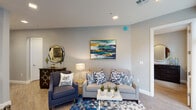 This well-appointed single-floor residence showcases a cohesive design aesthetic throughout, anchored by soft gray walls, light wood-look flooring, and a consistent blue-and-neutral color palette. The living area serves as a welcoming focal point, featuring a plush sofa accented with patterned throw pillows, a navy accent chair, and a bold teal-and-gold abstract painting that draws the eye upward. Recessed lighting and a geometric mirror add to the room's polished character.

The kitchen is a standout feature, outfitted with rich dark-wood shaker cabinetry extending to the ceiling, white quartz countertops, and a full suite of stainless steel appliances including a range, built-in microwave, dishwasher, and refrigerator. Ample upper and lower cabinetry ensures generous storage throughout.

The bedroom offers a serene retreat with a large upholstered headboard, dark wood dressers, a wall-mounted flat-screen television, and a window fitted with vertical blinds that allow natural light to filter in softly.

The bathroom features a backlit vanity mirror above a white quartz countertop, a dark wood vanity cabinet, and tasteful wall art, creating a clean and contemporary finish that complements the overall design of the space.