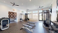 Located at 6000 Eldorado Pkwy in Frisco, TX, this fitness center offers a thoughtfully designed workout environment spread across a single, generously sized floor. The space is divided into two functional zones, each catering to different aspects of physical training.

The main workout area features durable rubber flooring throughout and is bathed in natural light from a series of large windows and transom windows positioned along the walls. Multiple ceiling fans keep the space comfortable, complemented by recessed lighting for consistent illumination. The cardio zone is lined with treadmills, ellipticals, and a stationary bike, all positioned to take advantage of the outdoor views. The strength training area includes adjustable benches, a TKO multi-functional cable machine, a lat pulldown station, a full dumbbell rack, and a rowing machine, offering a comprehensive range of equipment for all fitness levels. A large wall-mounted mirror and flat-screen TV enhance the training experience.

A secondary entry vestibule features a striking geometric-patterned accent wall and built-in cubbies, providing convenient storage for personal belongings. French-style glass doors open directly to the exterior, reinforcing the connection to the outdoors. The overall aesthetic is clean and modern, with crisp white walls and a polished, welcoming ambiance ideal for an active lifestyle.