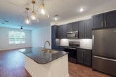 This single-floor residence showcases a thoughtfully designed open-concept layout anchored by a standout kitchen. The kitchen is equipped with tall, dark navy shaker-style cabinetry accented by gold bar pulls, providing generous storage that extends to the ceiling. A central island features a dark granite countertop with an undermount sink and a brushed gold faucet, illuminated by a trio of clear glass pendant lights with gold hardware. A herringbone-patterned tile backsplash frames the cooking area, which includes stainless steel appliances — a range, over-the-range microwave, and refrigerator. The kitchen flows seamlessly into an open living area finished with warm hardwood-style flooring throughout.

The bathroom offers a clean, functional design with large-format beige wall tile surrounding a soaking tub and shower combination fitted with gold fixtures and a matte black curtain rod. Built-in open shelving lines one wall, providing practical storage, while a dark granite vanity top with undermount sink mirrors the kitchen's design language.

The bedroom is a calm, carpeted retreat with neutral walls and a large window that allows natural light and greenery views to fill the space, creating a serene atmosphere throughout.
